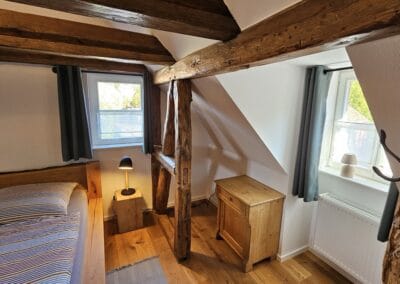 Gemütliches Schlafzimmer mit rustikalen Holzbalken, zwei Fenster mit Blick ins Grüne, moderner Einrichtung, Holzfußboden, entspannte Atmosphäre, ideal für erholsamen Aufenthalt.
